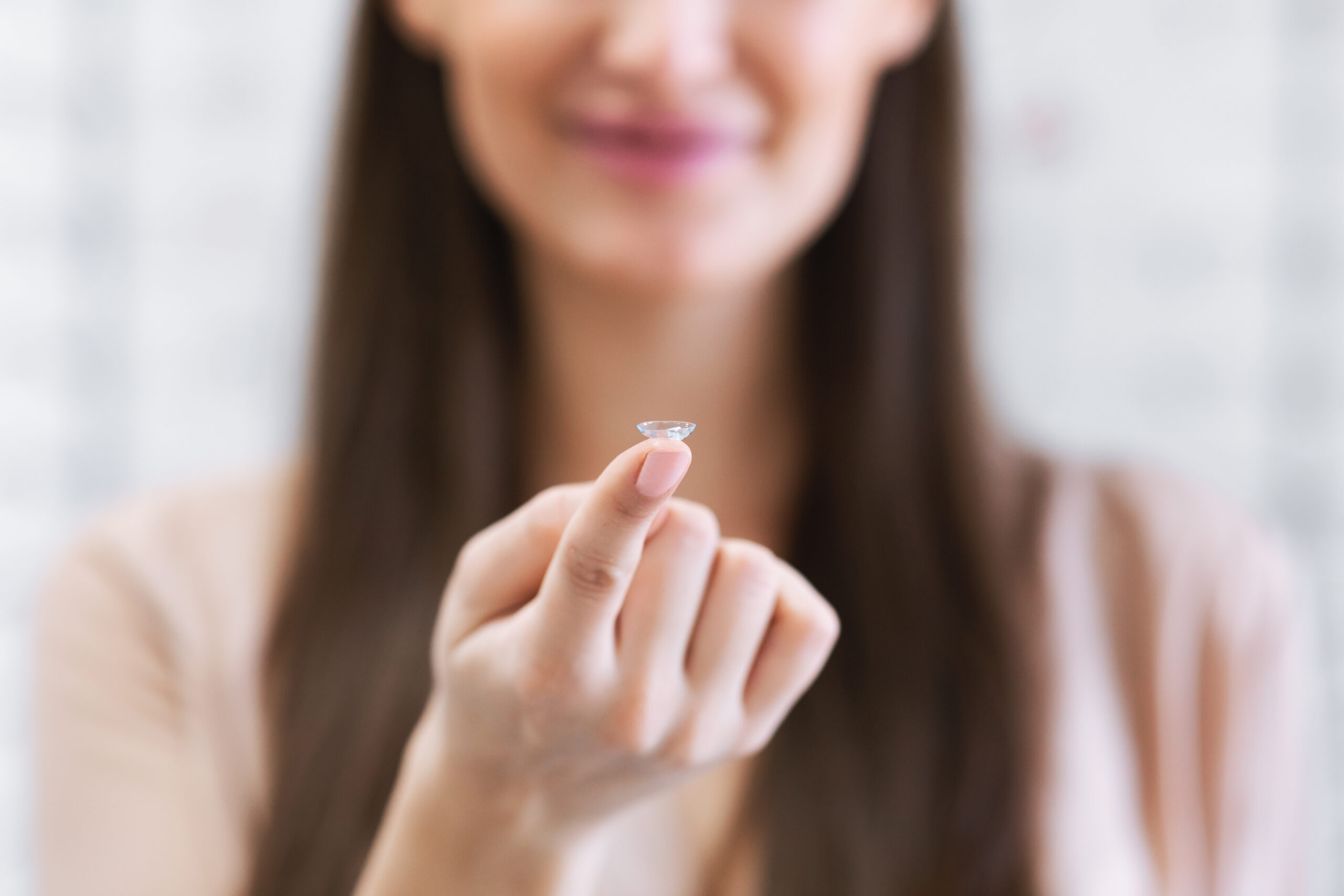 Optometry and Ophthalmology Concept. Closeup of unrecognizable smiling lady holding contact eye lens on finger and showing it to camera, selective focus, blurred background. Health and eye care